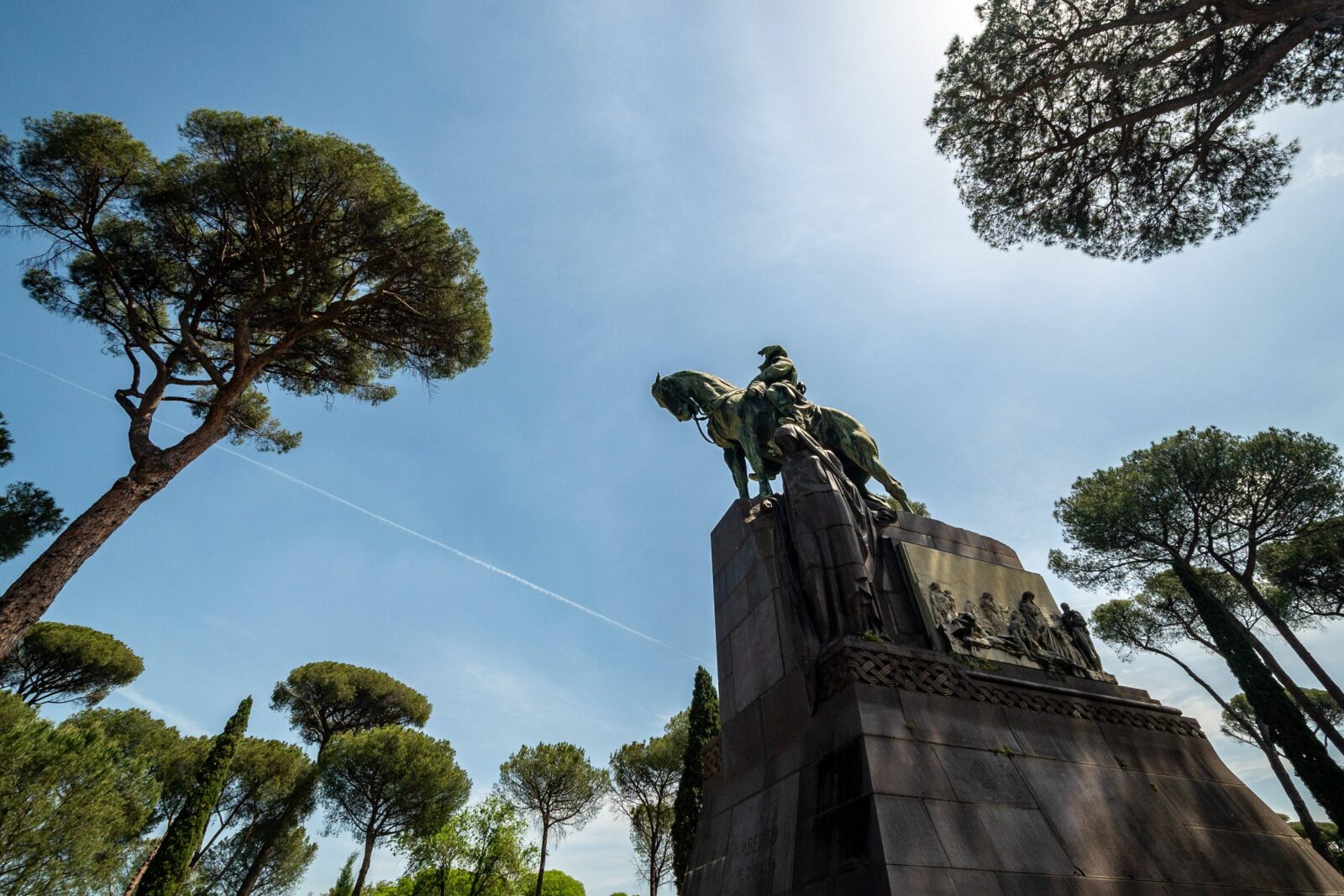 An ultrawide, low-angle shot from the left captures the Monumento Ad Umberto I, its architectural details visible against a sky partially obscured by tree branches extending inward from the top.
