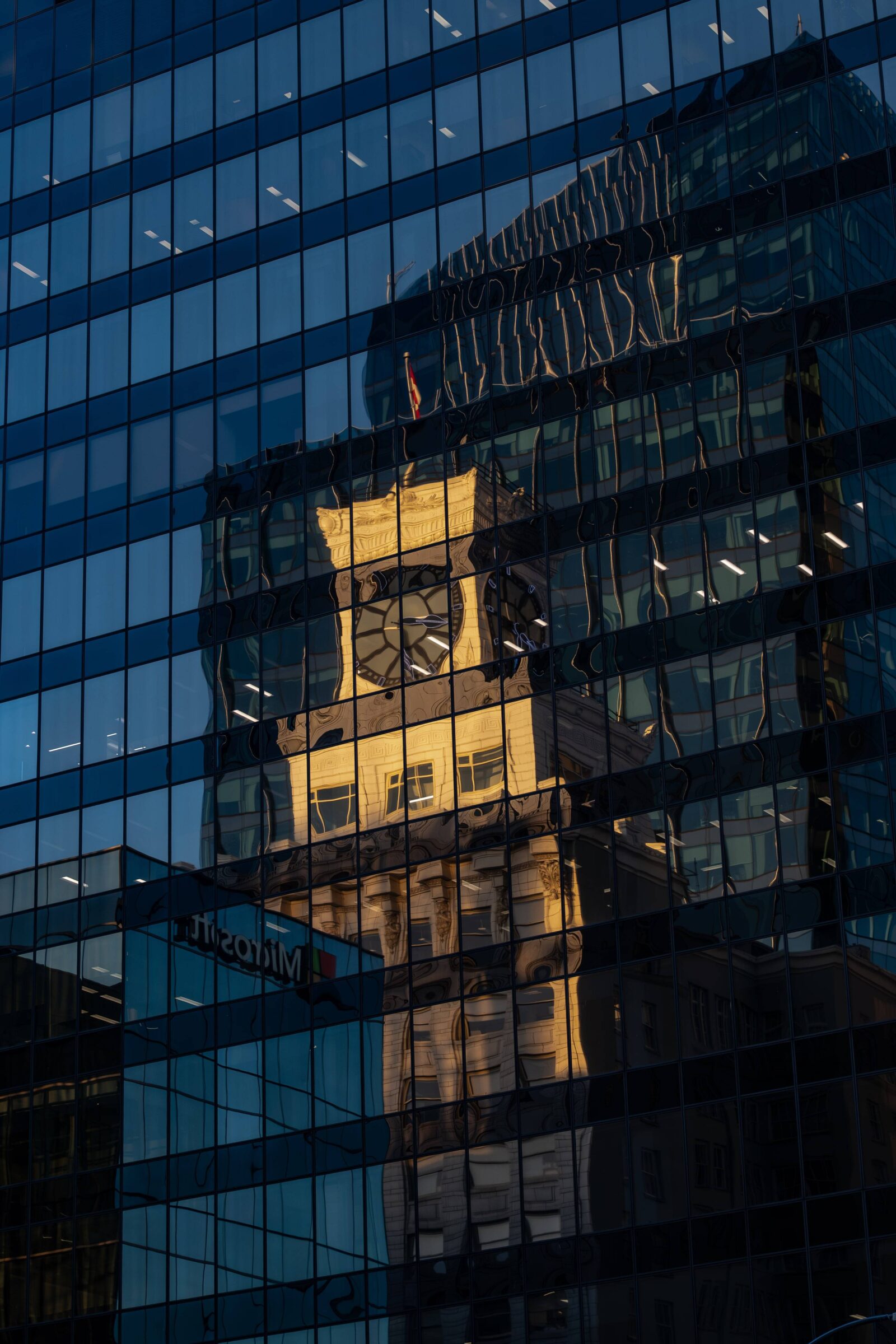 The golden-lit, classical architecture of the Vancouver Block building's upper floors and prominent clock tower are reflected in the deep blue, reflective windows of a contemporary glass office building. The reflection creates an interesting juxtaposition of old and new architecture in Vancouver's urban landscape.
