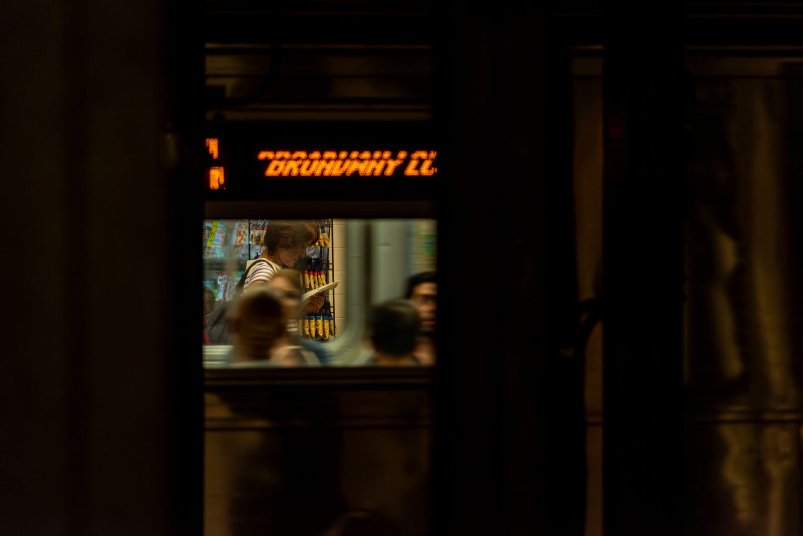 A passing NYC Subway train frames a distant lady across the gap reading a book.