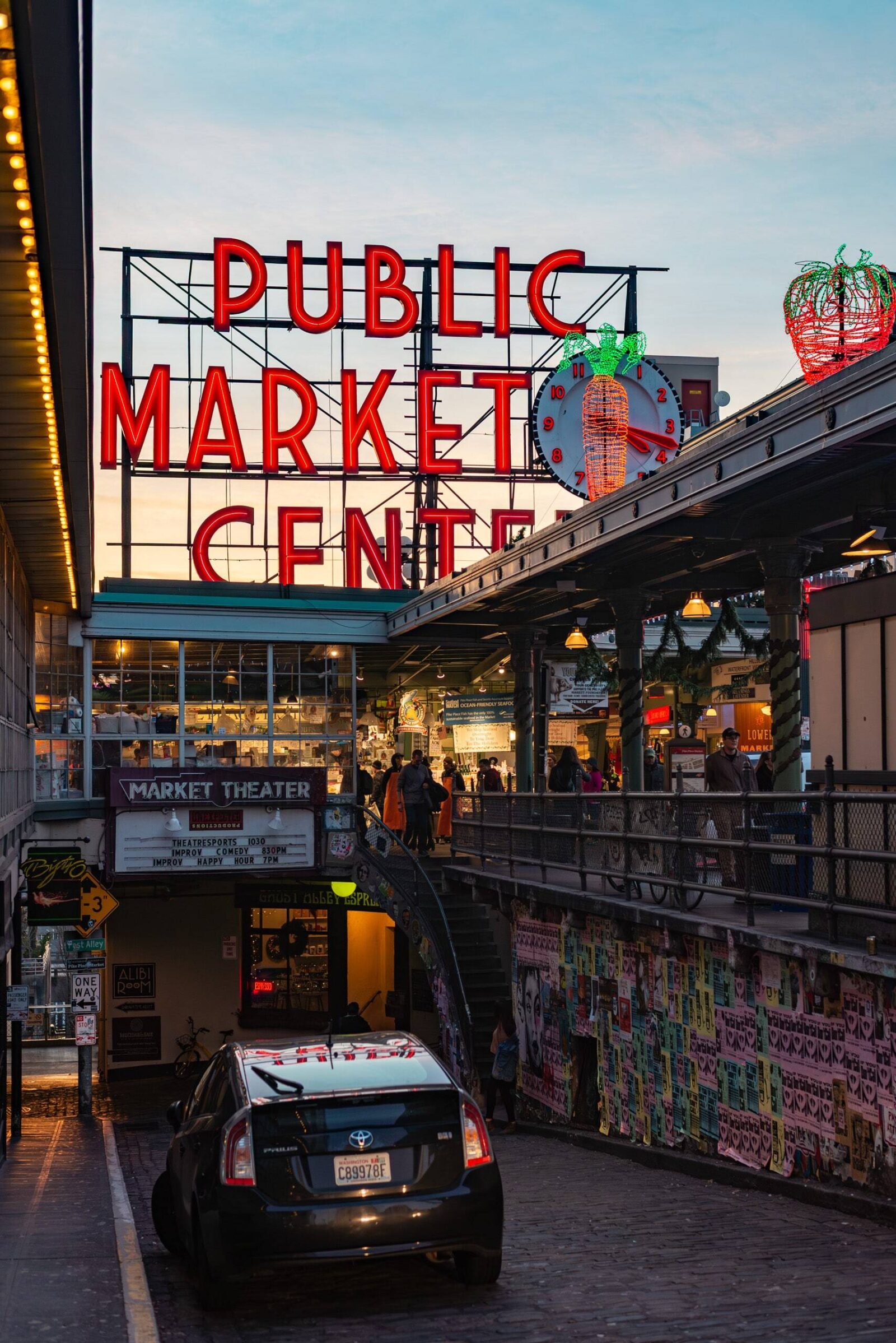 The famous Pike Place Market Center entrance with its red neon sign and clock, showing the bustling market activity below as visitors browse shops during the golden hour, with a car parked on the cobblestone street.