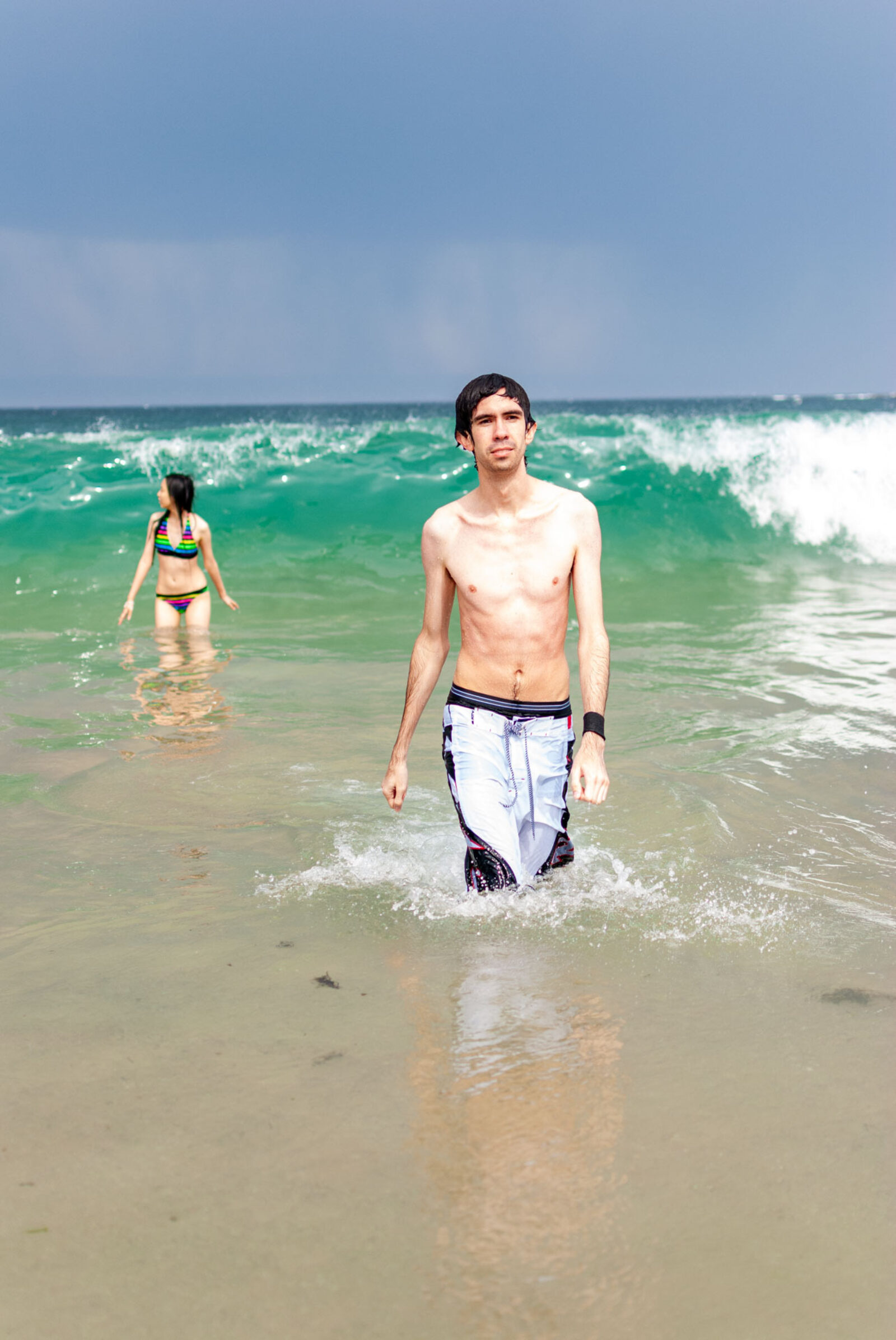 Image depicts a man walking casually as a lady at the back is about to get hit by a wave. The waves are as tall as the lady. Summer yet cloudy with peeks of sun lighting up the subjects as the sky behind displays a torrent of rain.