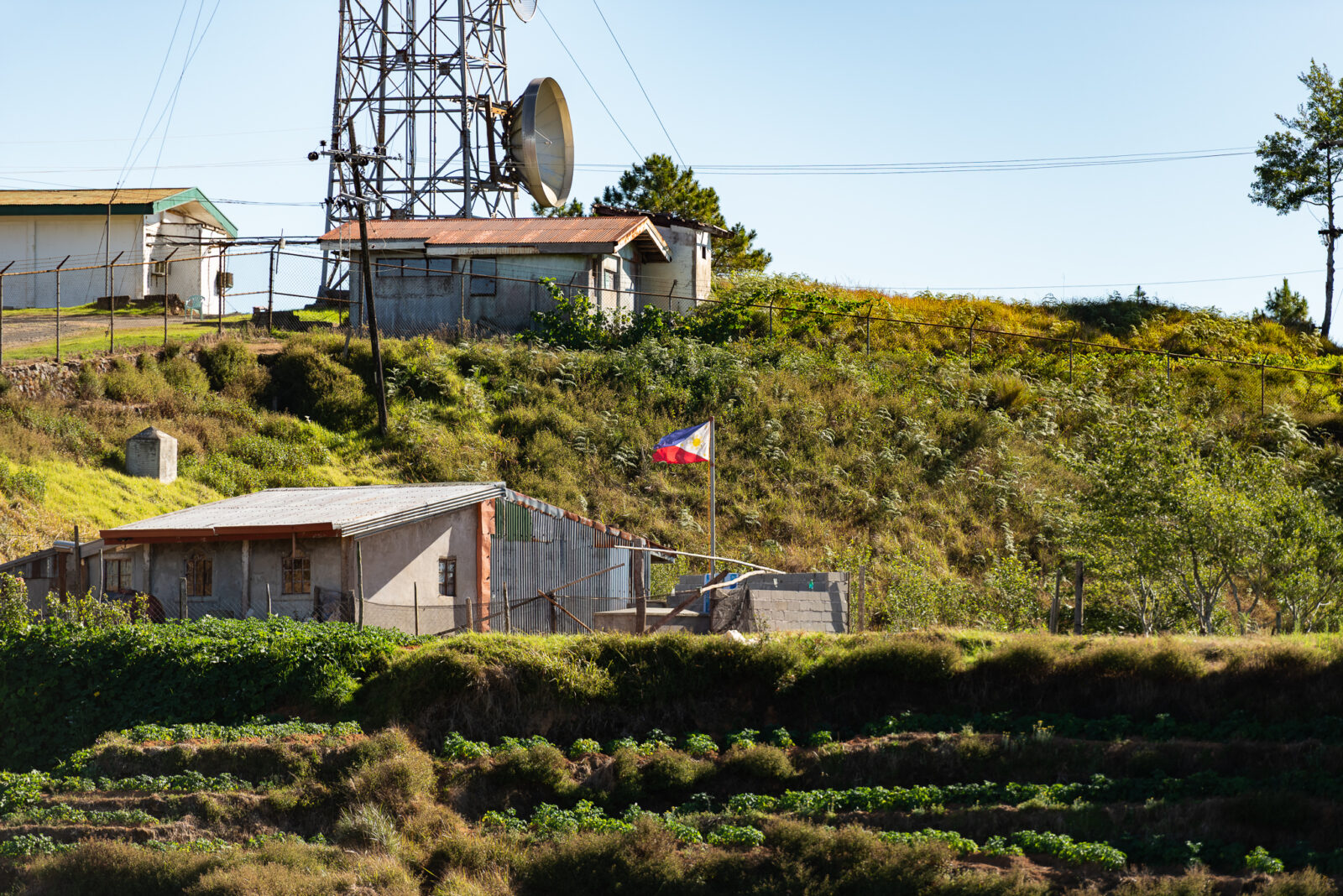 A rural hillside in Benguet with scattered small homes, terraced fields, a tall communication tower, and a Philippine flag waving.